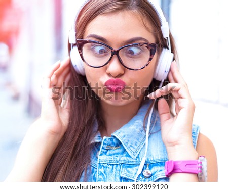 Close up funny portrait of funny young pretty girl listening music and going crazy, wearing jeans vest,white headphones and trendy clear sunglasses, posing at city center at sunny summer morning day.