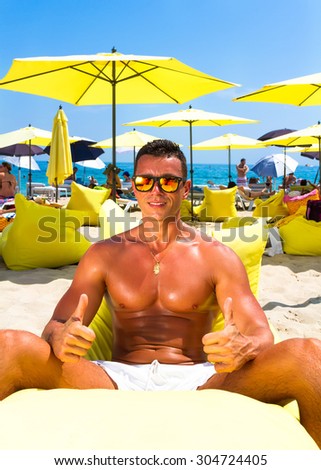 Portrait of a handsome young muscular man in white swimtrunks with luxury beach resort background,sitting on beach chair and showing super sign,getting fun on summer vacation