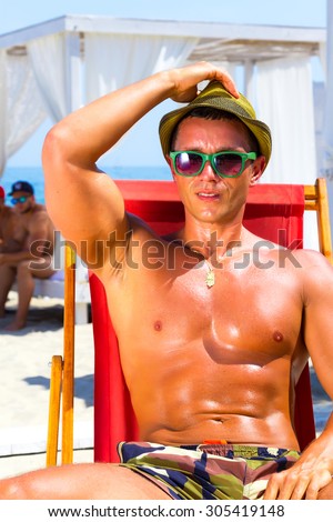 Sexy hot muscular Man on beach looking to camera smiling ,wearing hipster hat, sunglasses,military printed trousers.Young male model enjoying summer travel holiday by the ocean on luxury beach resort
