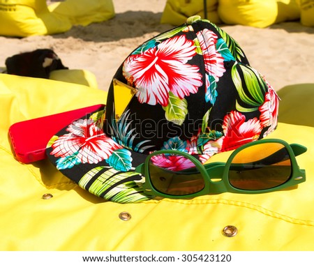 Travel, summer vacation, tourism and objects concept. close up of floral printed hat, hipster green sunglasses,smartphone in red cover on yellow beach chair.articles for beach vacations.