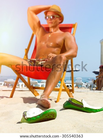 Sexy hot muscular Man on beach looking to camera smiling ,wearing hipster hat, sunglasses,military printed trousers.Young male model enjoying beach with shoes off.Focus on shoes.