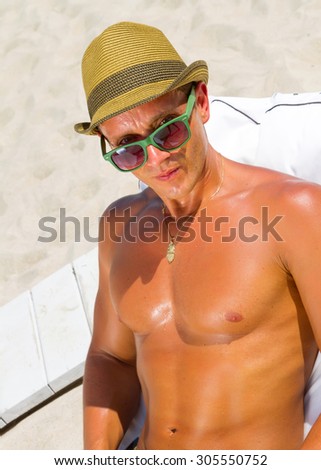 Sexy hot muscular Man on beach looking to camera ,wearing hipster hat, sunglasses.Young male model enjoying summer travel holiday by the ocean on luxury beach resort