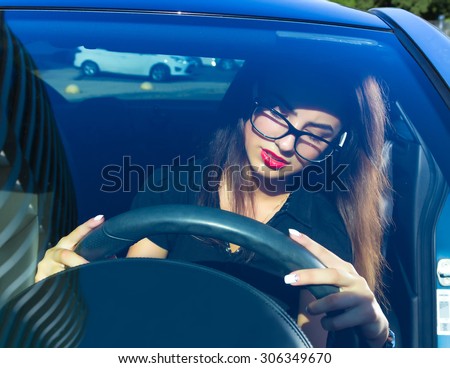 Young woman driving her car.Stylish student girl in car.Summer fashion close up portrait of elegant beautiful woman driving her car in urban city, wearing black shirt and black clear glasses.
