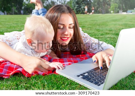 Young mother and baby boy looking into laptop.Happy brunette mother with her baby boy using laptop in the park.Mother and adorable baby play with laptop.Happy family, mother and son resting in park
