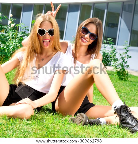 Attractive best friends having fun in city park. Hipster Girls Laughing and having fun on summer holidays and vacation-girls going crazy on the grass.Wearing similar outfit.Fun,joy,playful mood.