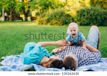 Young happy family of three lying on blanket and having fun in the park. Pretty woman with husband and cute little baby boy. Father holding his son at arm\'s length. Happy parenting concept.