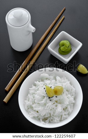 A dish of steamed rice with chopstick, soy sauce jug and wasabi paste and asian pickles on a black background