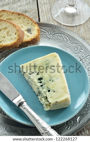 Blue cheese slice on a grey blue plate with baguette slices,knife and base of a wine glass above shot