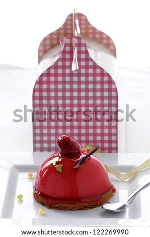 A raspberry mousse pÃ?Â¢tisserie / pastry with a red glaze, fresh raspberry and gold leaf and chocolate decoration with a pink chequered pastry box in the background