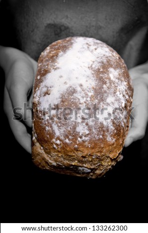 A multi grain loaf of bread dusted with flour being hald in two hands. The bread is in color and the hands are black and white.