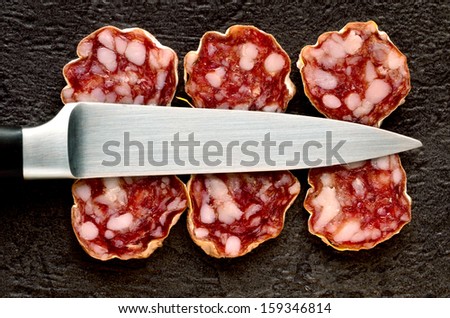 Saucisson slices on a black background with a stainless steel knife resting on top.