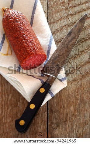 A salami sausage on a tea towel with an old carbon steel french knife at an angle and wooden board