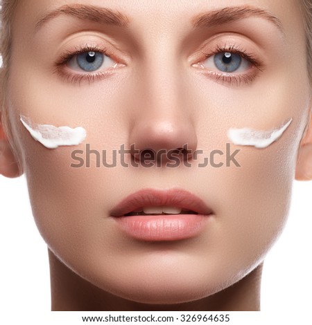 Beautiful face of young woman with cosmetic cream on a cheek. Skin care concept. Closeup portrait isolated on white. Close-up young beautiful face of girl applying moisturize cream.