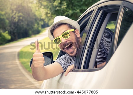 Young man wearing hat and sunglasses showing thumbs up from driverâ??s seat through opened window. Vacation and travel concepts.