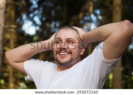Portrait of a smiling surprised man with beard standing and looking ahead against a green trees in dark forest with hands up