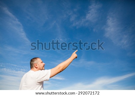 Side portrait low angle view of a man with beard standing and looking ahead against a blue sky with white clouds and pointing with finger