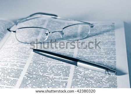 open dictionary with a pair of glasses and pen laying on top. Blue toning