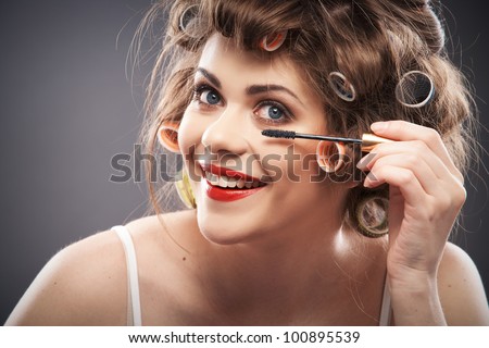 Close up portrait of a young woman with long hair on gray background making beauty face and hair style, applying mascara. Smile happy girl  with make up accessories, studio isolated.