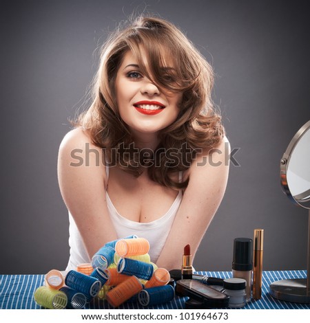 Portrait of a young woman with long hair on gray background isolated.  Happy girl seating at table with make up accessories and mirror. Smiling model with curler hair dress