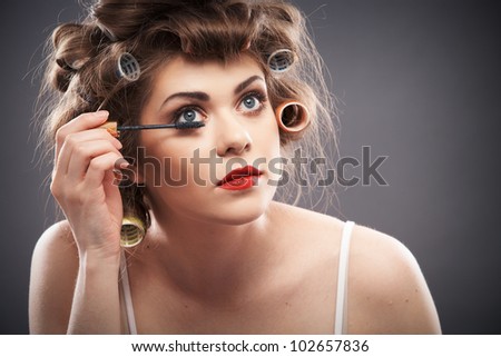 Close up portrait of a young woman with long hair on gray background making beauty face and hair style, applying mascara. Smile happy girl  with make up accessories, studio isolated.