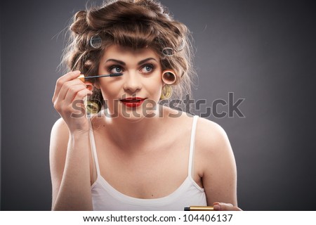 Close up portrait of a young woman with long hair on gray background making beauty face and hair style, applying mascara. Smile happy girl  with make up accessories, studio isolated.