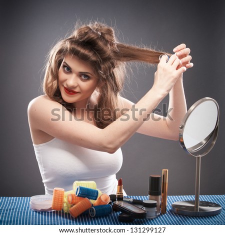 Portrait of a young woman with long hair on gray background isolated.  Happy girl seating at table with make up accessories and mirror. Smiling model with curler hair dress beauty style portrait