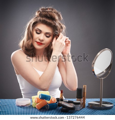 Portrait of a young woman with long hair on gray background isolated.  Happy girl seating at table with make up accessories and mirror. Smiling model with curler hair dress beauty style portrait