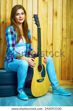 Girl hold guitar, sitting on a car  wheels against wooden background.