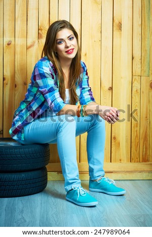 Girl in country style sitting on auto tires against yellow wooden wall. Smiling model with long hair.