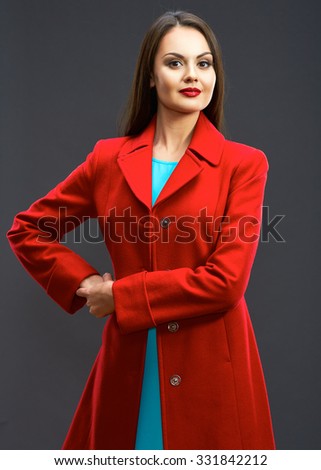 Smiling fashion model with long hair red coat dressed standing against gray studio background, isolated.
