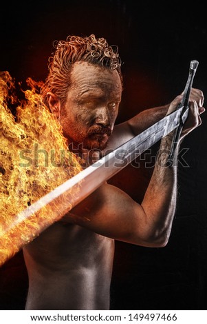 Strong warrior licking a big sword in fire, covered in mud and naked