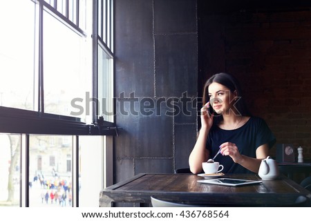 Young attractive girl talking on mobile phone and smiling while sitting alone in coffee shop during free time. Happy female having rest in cafe. Lifestyle