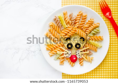 Lion pasta - fun food idea for kids lunch, animal shaped food art. Colorful fusilli vegetables pasta with sandwich like a cute lion head on white plate top view