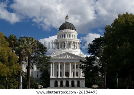 an image of the California State Capital Building from a distance bordered by trees and a blue sky with grey and white clouds