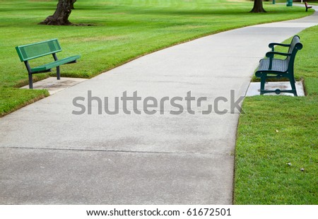 Two green metal benches across a large concrete pathway in a park