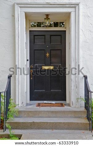 Black front door to white house with newspaper and lamp