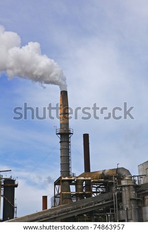 Old factory smoke stack billowing a white gray plume across blue and white cloud sky