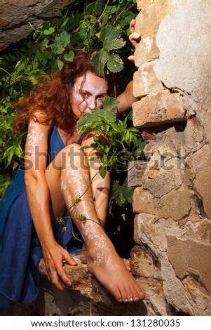 Woman with white body henna tattoos hiding in bush behind brick wall.