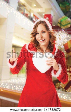 happy smiling Santa woman indoors, holding white heart, thumb up, background digitally added, work path