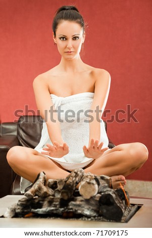 Young female sitting in lotus position, warming her arms on modern fireplace