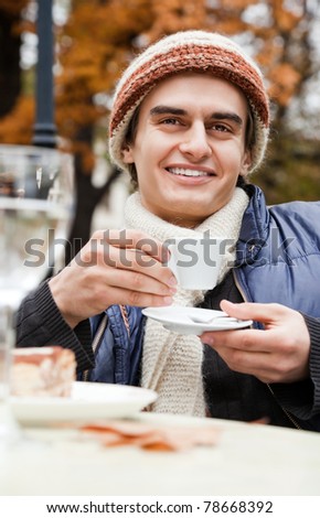Young happy man wearing hat sitting and drinking coffee in park on autumn day