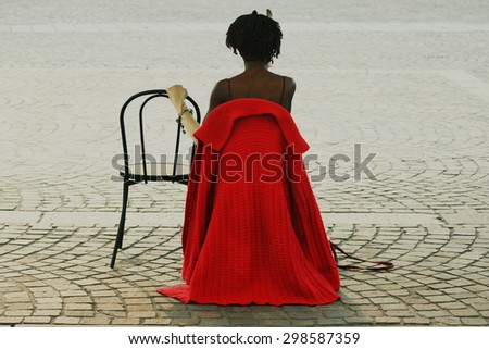 ITALY - Black woman wears a long red knitted cardigan and a white glove while sitting on a chair in Piazza San Carlo in Turin