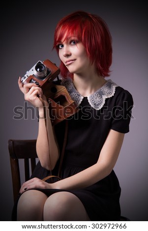 Red-haired girl, vintage camera, piercings on his face, wooden chair, taking a picture, enigmatic smile, dress with lace, black nail polish, a mystical image, blue eyes, informal appearance.