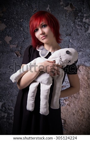 Red-haired girl, gloomy story, rag doll, piercings on his face, a mystical image, cracked plaster, blue eyes, portrait on texture background, girl with a doll, informal appearance, handmade doll.