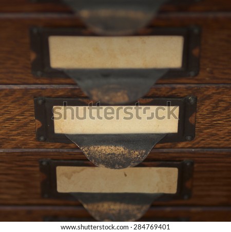 Stack of three old, oak flat file drawers with blank yellowed tags in tarnished brass label holders. Shallow DOF with focus and lighting on the middle drawer.