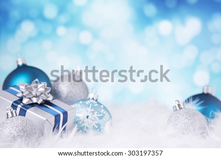 Blue and silver Christmas baubles and a gift on a soft feathery surface in front of defocused blue and white lights.