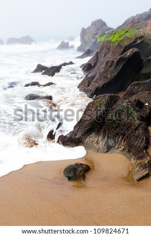Beach with boulders in the mist and fog.  The tide is rushing in with foam on the sand.  Location is San Francisco near the foot of the Golden Gate Bridge.  Copy space
