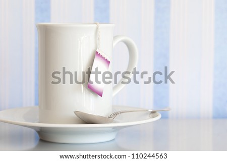 White cup and teabag with a blank label, and a sterling silver spoon on a reflective surface.  Blue and white striped background.