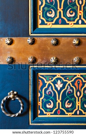 Moroccan Wooden Painted Door. Close Up Detail Of Floral Design On A Deep Blue Background, Bronze Knocker And Metal Nail Heads. Location: Marrakech, Morocco
