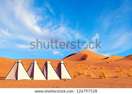White canvas tents in a Sahara desert camp. Colorful image with orange sand dunes and blue sky with swirling clouds.